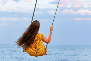 Beautiful girl in yellow dress with long hair is riding on swing above sea. Back view