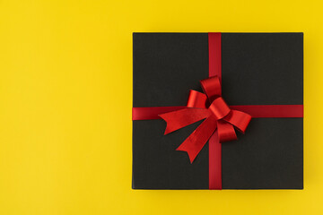 Black box with red ribbon and bow on bright yellow background. Top view