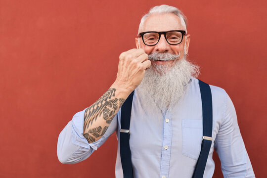 Happy hipster senior man touching mustache with red wall in background - Focus on face