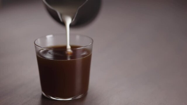 Slow motion milk pour into tumbler glass with coffee on walnut table