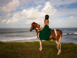 Woman riding horse near the ocean. Outdoor activities. Asia woman wearing long green dress. Traveling concept. Cloudy sky. View from back. Copy space. Bali