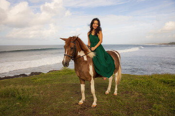 Happy woman riding horse near the ocean. Outdoor activities. Asia woman wearing long green dress....