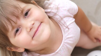 Close-up portrait of smiling cute little girl looking at camera and shaking head with long blond hair, hazel eyes, handheld high angle view. Top view of cheerful laughing child having fun, summer day