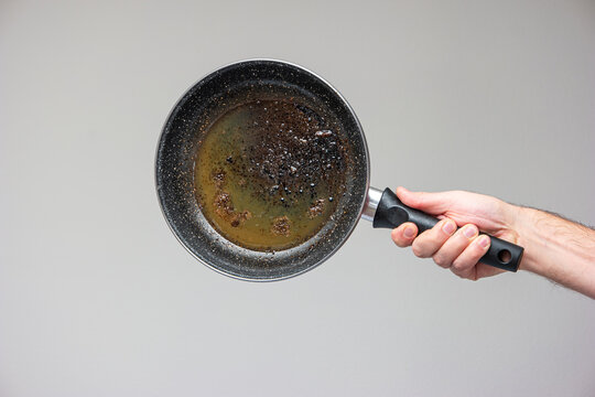 Caucasian male hand holding an old frying pan stained with brown burned oil and grease isolated on gray