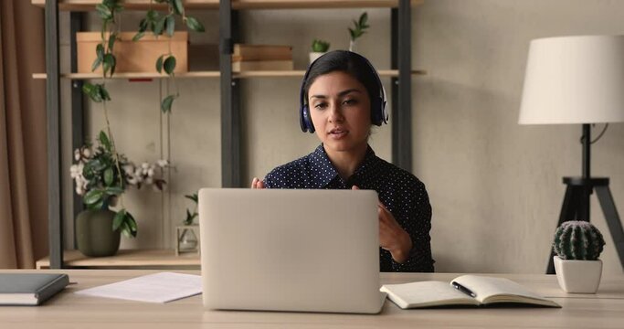 Focused young indian ethnicity business lady wearing wireless headphones, looking at laptop screen, involved in web camera video call conversation with colleagues or giving educational online class.