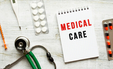 MEDICAL CARE is written in a notebook on a white table next to pills and a stethoscope.