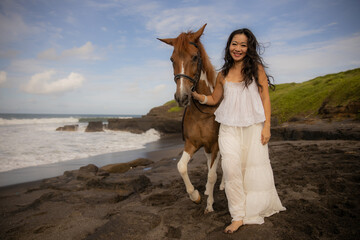Smiling woman leading horse by its reins. Horse riding on the beach. Human and animals relationship. Asian woman wearing long white dress. Nature concept. Copy space. Bali