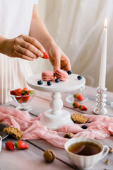 Female hands put strawberries on a wooden white cake stand. Exquisite serving of macaroon dessert with berries.