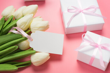 lose up view of white tulips and gift boxes on pink background with blank gift tag. Mother's day or women's day composition.
