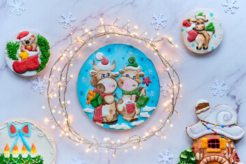 Decorated gingerbread with two cartoon cow characters, wintertime house, Santa and Xmas wreath. Christmas flat lay with festive garland wrapped as wreath. Top view on white marble stone background.