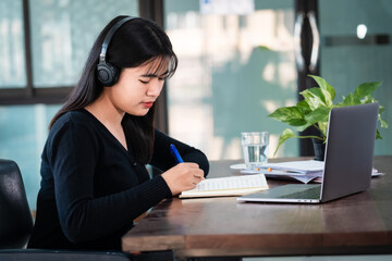 Smiling Asian girl student wears wireless headphones write on the notebook to study language online watch and listen to the lecturer, webinar via video call e-learning at home, distance education