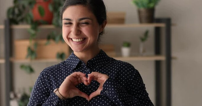 Happy millennial kind indian ethnicity woman showing heart symbol with fingers, feeling thankful. Smiling compassionate mixed race female volunteer demonstrating support and care sign to camera.
