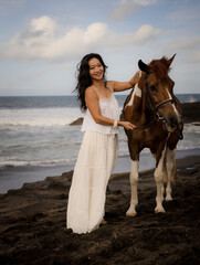 Woman leading horse by its reins. Horse riding on the beach. Human and animals relationship. Asian woman wearing long white dress. Travel concept. Copy space. Bali