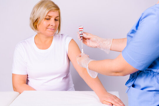 Senior Blonde Woman Awaiting Coronavirus Vaccine. Nurse Hands Prepare Syringe And Ampoule Of Covid 19 Vaccine.