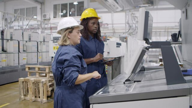 Middle aged female factory worker taking proficiency exam, explaining how to operate new machine to black mentor at control panel. Industry or machinery concept