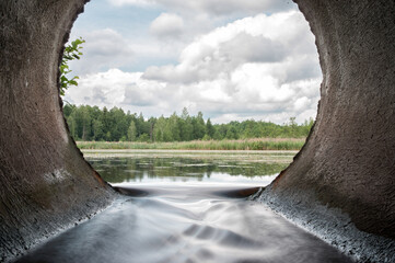 Perfect view from concrete drain pipe in the forest river. Beautiful water flow from the lake. - obrazy, fototapety, plakaty
