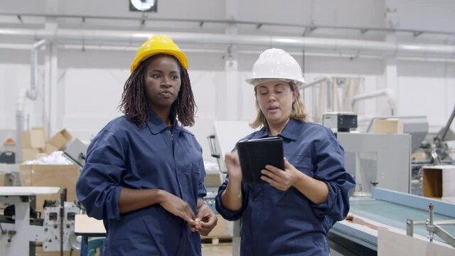 Factory mentor training intern. Diverse colleagues in protective uniforms using tablet, talking, discussing machines and equipment work on plant floor. Dolly shot. Industrial occupation concept