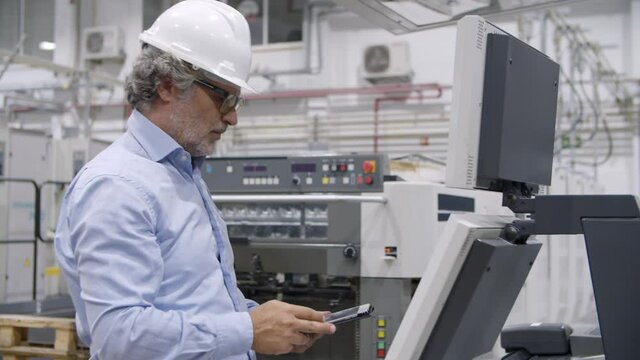 Older factory engineer testing industrial machine, pressing buttons at control board, monitoring process, using tablet. Production process or machinery concept