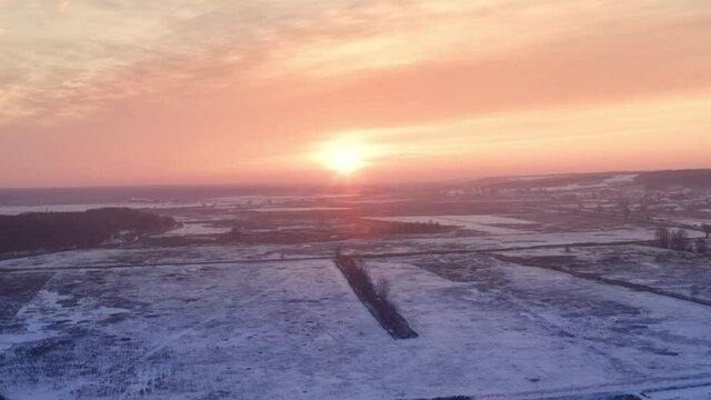 Bright sun in the clear sunset evening sky in frosty weather - golden hour. A drone flight over a snow-covered rural landscape to the setting sun on the horizon.