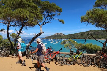 Randonneurs marchant sur un sentier de randonnée de l'île de Porquerolles, et vélos garés devant la baie et la plage Notre-Dame, au bord de la mer Méditerranée (France) - obrazy, fototapety, plakaty