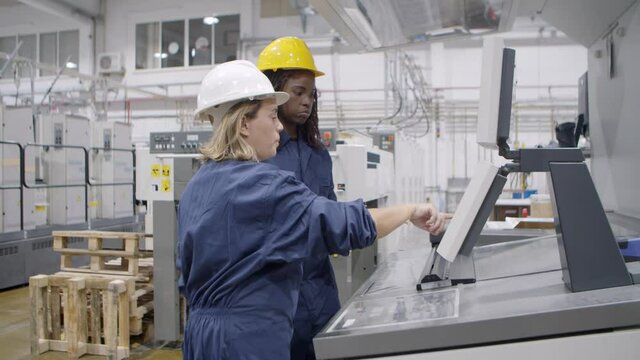 Middle aged female factory employee taking proficiency exam, explaining how to operate new machine to black mentor at control panel. Industry or machinery concept