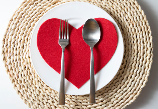 St Valentine's table setting with festive decorations on a wicker stand, with white plate, a fork with a tablespoon with red heart on it 