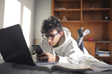 young boy studies lying on the bed using computer and smartphone - 409484687