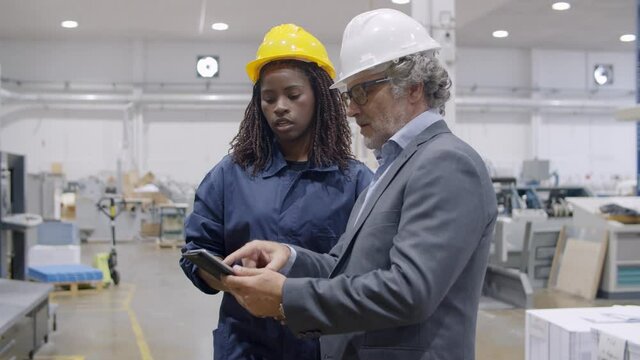 Factory mentor training Black female employee or intern. Diverse colleagues in hardhats talking on plant floor, man using tablet. Static shot. Industrial occupation concept