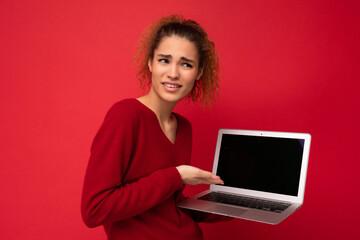 Close-up portrait of beautiful amazed surprised astonished dark blond woman holding laptop computer looking to the side showing at netbook keyboard wearing red sweater isolated over red wall