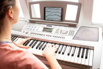 Piano online courses. Grirl is practicing playing on electric piano using tablet standing on the desk