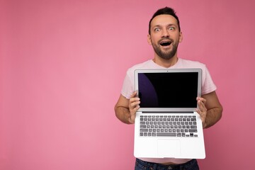 Handsome shoked man holding laptop computer looking at camera in t-shirt on isolated pink background