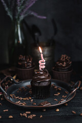 Several muffins or cupcakes with chocolate shaped cream at black table. Festive candle burns on a chocolate cake.