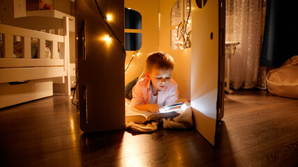Happy smiling boy lying on floor at hos toy house or tent and reading with flashlight at night....