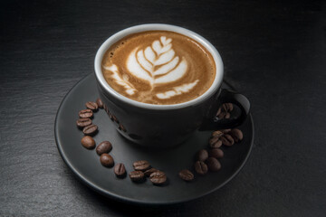 coffee in black cup with leaf image on foam and with coffee beans on saucer, black slate stone background