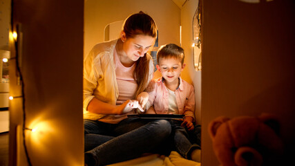 Little boy with mother playing in toy cardboard house and using tablet computer. Concept of child education and family having time together at night