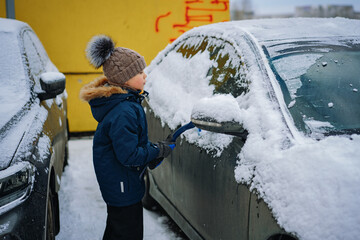 cute little caucasian boy wearing  knit hat and hooded jacket brushing snow from a car on winter...