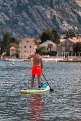 A young athletic man stands on a SUP stand up paddle board in Kotor Bay overlooking a beautiful...