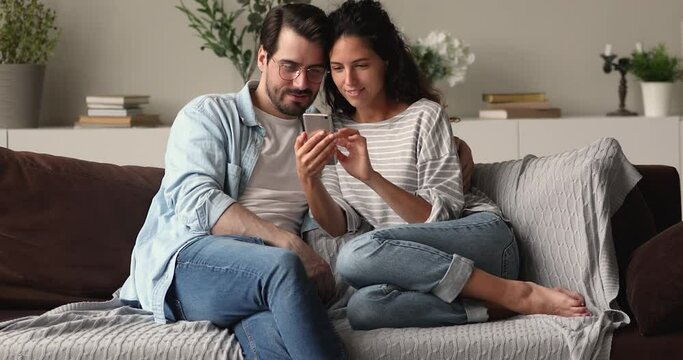 Millennial couple in love relaxing on couch at home use smartphone enjoy weekend choose goods, buy on internet, discuss purchase, read social media news. Modern tech usage, free time, leisure concept