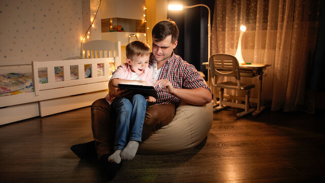 Happy laughing boy with young father sitting in bedroom at night and playing games on tablet computer. Concept of child education and family having time together at night.