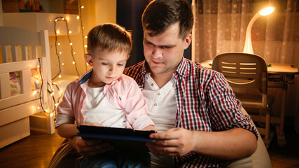 Smiling boy with father using tablet computer in bedroom decorated for Christmas. Concept of child education and family having time together at night.