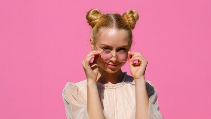 Portrait of happy blonde teenager girl with sunglasses, smiling, looking out from under the glasses on pink background in summer. Positive Emotions. Lifestyle. 