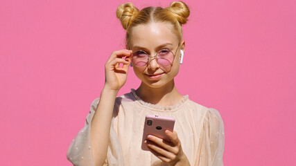 Young happy blonde girl teen blogger makes selfie on smartphone in wireless headphones, in pink...