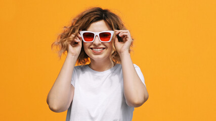 Portrait of happy pretty woman stylish white sunglasses with red glasses raises up, smiling with pleasure on yellow background in summer. Emotions. Positive girl. Lifestyle. Freedom