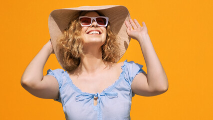 Portrait of happy pretty woman wearing hat and in stylish sunglasses, smiling, looking at sun with pleasure, adjusting her pink hat on yellow summer background. Emotions, happiness, joy. Positive girl