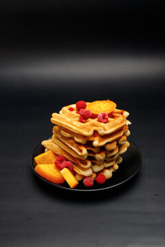 Luxurious traditional waffle pile with ripe raspberries, blueberries and syrup dropping isolated on black background. Culinary sweet dessert.