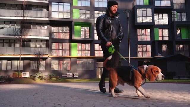 Handsome man in leather jacket walking with his dog on street. Young guy holding his lovely pet on leash outdoors. Best friends at urban area.