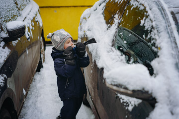 cute little caucasian boy wearing scandinavian knit hat  brushing snow from a car on winter day . Image with selective focus