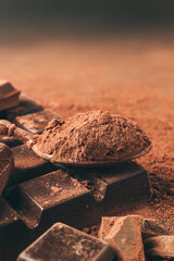 Brown cocoa powder in the spoon and chopped chocolate cubes, on dark background with copy space