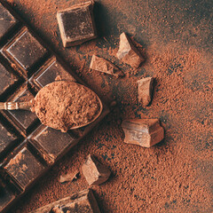 Brown cocoa powder in the spoon on chocolate bar and chopped chocolate cubes, top view on dark background