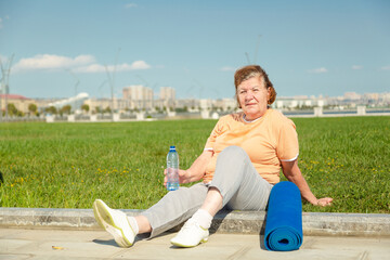 Pensioner woman resting after training in the park. Fitness training for aged people. Healthy...